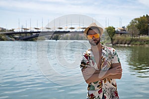 Young and handsome man, mirror sunglasses, beard, hat, Hawaiian shirt and jeans on a pier by the river, arms folded. Concept