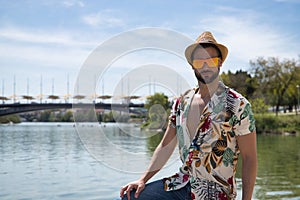 Young and handsome man, mirror sunglasses, beard, hat, Hawaiian shirt and jeans on a pier by the river. Concept vacation, party,