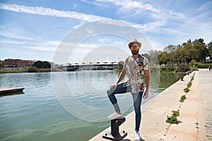 Young and handsome man, mirror sunglasses, beard, hat, Hawaiian shirt and jeans on a pier by the river. Concept vacation, party,