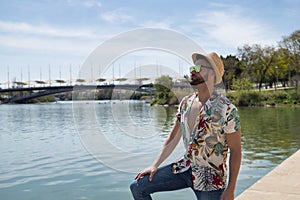 Young and handsome man, mirror sunglasses, beard, hat, Hawaiian shirt and jeans on a pier by the river. Concept vacation, party,