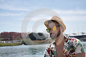 Young and handsome man, mirror sunglasses, beard, hat and Hawaiian shirt, sitting on a pier by the river. Concept vacation, party