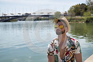 Young and handsome man, mirror sunglasses, beard, hat and Hawaiian shirt, sitting on a pier by the river. Concept vacation, party