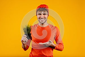 Young handsome man in orange hat holds grass houseplant, isolated yellow background