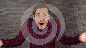 A young handsome man in plaid shirt opens his mouth and spreads his hands from surprise on a gray brick background.