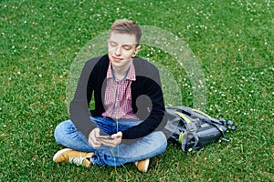 Young handsome man in plaid shirt sitting on green lawn with closed eyes and  listening to music with headphones from smart phone