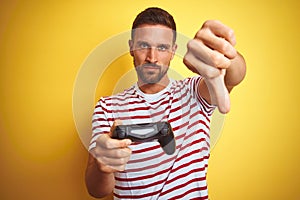 Young handsome man playing video games using joystick game pad over yellow background with angry face, negative sign showing