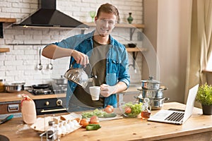 Young handsome man pouring hot water from the kettle making tea standing in well-equipped modern kitchen checking recipe in laptop