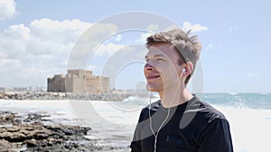 Young handsome man puts the earphones on before run workout standing on the beach smiling. Castle on the background