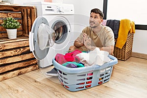 Young handsome man putting dirty laundry into washing machine begging and praying with hands together with hope expression on face