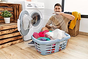Young handsome man putting dirty laundry into washing machine gesturing with hands showing big and large size sign, measure symbol