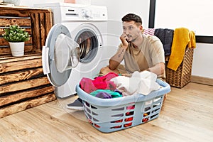 Young handsome man putting dirty laundry into washing machine looking stressed and nervous with hands on mouth biting nails