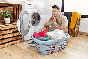 Young handsome man putting dirty laundry into washing machine praying with hands together asking for forgiveness smiling confident