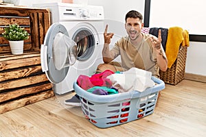 Young handsome man putting dirty laundry into washing machine shouting with crazy expression doing rock symbol with hands up