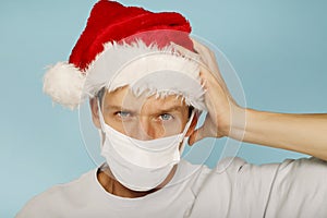 Young handsome man in red christmas santa hat and white health care face mask standing with hand on head over blue background