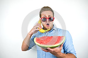 Young handsome man in red eyeglasses with watermelon holding banana like a phone on isolated white background