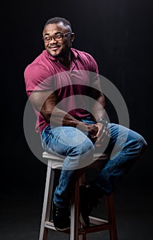 Young handsome man in red shirt and jeans sitting on chair. Athletic muscular male posing for camera on chair.