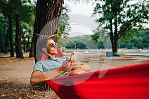 Young handsome man resting in the park lying on a hammock with a smartphone in his hands, looking away with a serious face