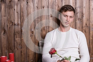 Young handsome man with a rose in his hands sitting near wood wall