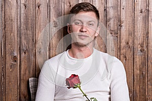 Young handsome man with a rose in his hands sitting near wood wall