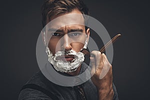 Young handsome man with shaving cream on his face, grooming his beard with straight razor and looking