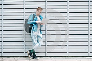 Young handsome man in shirt, jeans and glasses carrying a backpack using laptop computer standing on a wooden panel background.
