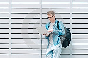 Young handsome man in shirt, jeans and glasses carrying a backpack using laptop computer standing on a wooden panel background.