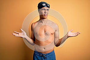 Young handsome man shirtless wearing swimsuit and swim cap over isolated yellow background clueless and confused with open arms,