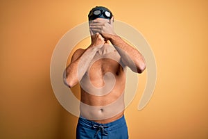 Young handsome man shirtless wearing swimsuit and swim cap over isolated yellow background Covering eyes and mouth with hands,