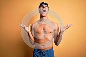 Young handsome man shirtless wearing swimsuit and swim cap over isolated yellow background crazy and mad shouting and yelling with