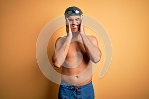 Young handsome man shirtless wearing swimsuit and swim cap over isolated yellow background with hand on headache because stress
