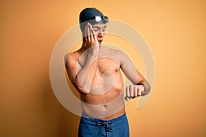 Young handsome man shirtless wearing swimsuit and swim cap over isolated yellow background Looking at the watch time worried,