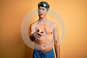 Young handsome man shirtless wearing swimsuit and swim cap over isolated yellow background pointing displeased and frustrated to