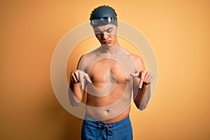 Young handsome man shirtless wearing swimsuit and swim cap over isolated yellow background Pointing down looking sad and upset,