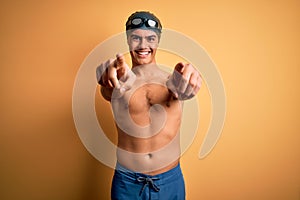 Young handsome man shirtless wearing swimsuit and swim cap over isolated yellow background pointing to you and the camera with