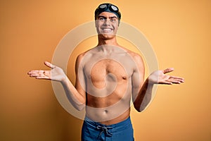 Young handsome man shirtless wearing swimsuit and swim cap over isolated yellow background smiling cheerful with open arms as