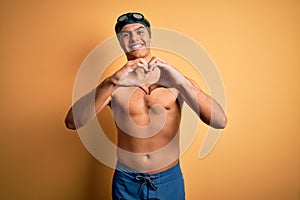 Young handsome man shirtless wearing swimsuit and swim cap over isolated yellow background smiling in love doing heart symbol