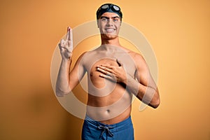 Young handsome man shirtless wearing swimsuit and swim cap over isolated yellow background smiling swearing with hand on chest and
