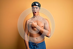 Young handsome man shirtless wearing swimsuit and swim cap over isolated yellow background Surprised pointing with finger to the