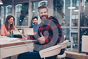 Young handsome man showing thumb up while his collegues working on project, sitting on modern workplace