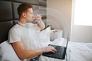 Young handsome man sit in bed this morning. He drink water. Male model hold laptop. Headphones around neck.