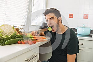 A young handsome man sits in the kitchen and reluctantly eats vegetables.