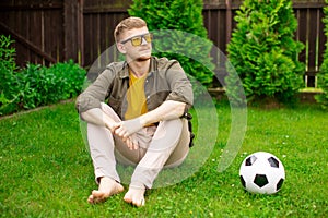 Young handsome man sits on lawn at home with soccer ball on grass, championship