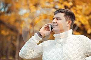 Young handsome man sitting in autumn park and talking by phone. Businessman is calling to someone by smartphone