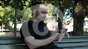 Young handsome man sitting on bench browsing internet via phone.