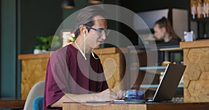 Young handsome man sitting in cafe with laptop making video calling using webcam and headphones