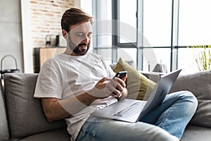 Young handsome man sitting on couch in living room at home using computer chatting and holding smartphone