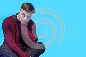 Young handsome man sitting on the floor on blue background.  Fashion hairstyle, expressive harsh look to the camera.