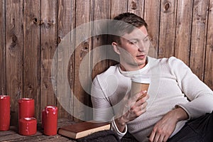 Young handsome man sitting near wood wall drinking coffee and reading book