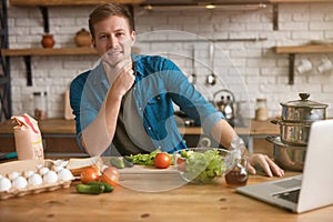 Young handsome man sitting in well-equipped kitchen checking recipe for tasty breakfast in his laptop , sunday morning