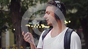 Young handsome man with smartphone in hand and headphones listening to music on green street of evening city